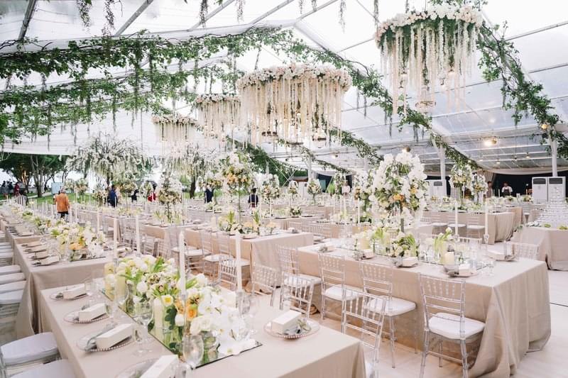  Luxurious wedding reception setup under a transparent marquee at Alila Villas Uluwatu, featuring long banquet tables with floral centerpieces, hanging floral chandeliers, and greenery accents 