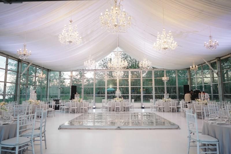  Elegant wedding reception setup in a glasshouse at Alila Villas Uluwatu, featuring white draped ceilings, sparkling chandeliers, round tables, and a mirrored dance floor. 