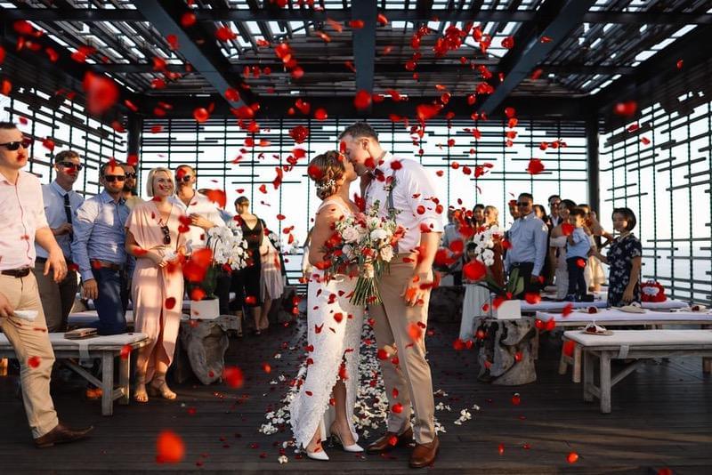  Bride and groom share a kiss surrounded by friends and family inside the cabana at Alila Villas Uluwatu, with vibrant red rose petals falling to celebrate the moment 