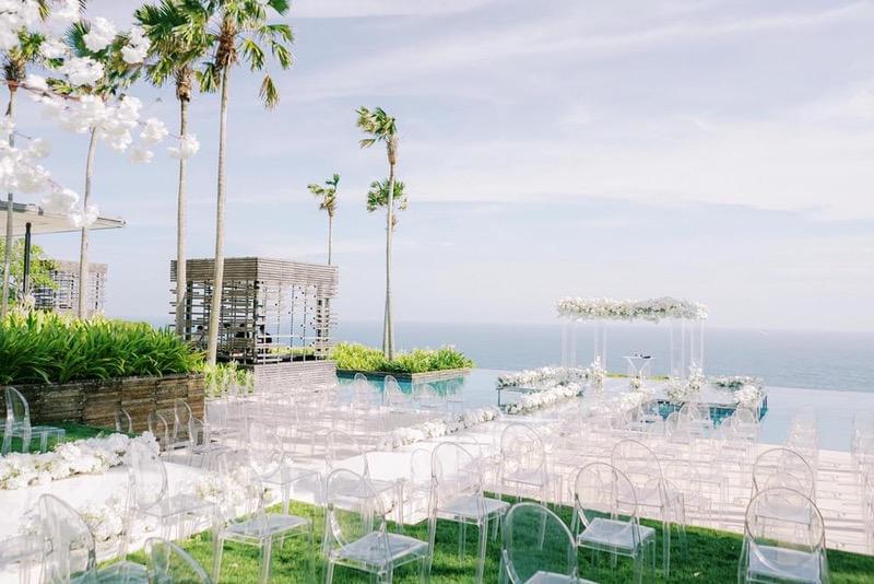  Wedding ceremony setup at Alila Villas Uluwatu, featuring clear acrylic chairs, a white floral aisle, and a floating altar with ocean views and tropical palm trees in the background. 