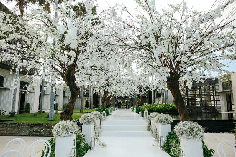  Wedding aisle at Alila Villas Uluwatu, lined with white floral arrangements and illuminated by lanterns, beneath arching artificial cherry blossom trees creating a romantic ambiance. 