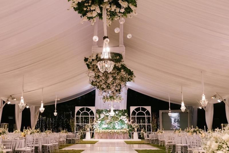  uxurious wedding reception setup at Alila Villas Uluwatu, featuring draped ceilings, crystal chandeliers adorned with floral arrangements, a grand stage backdrop, and elegant round tables with floral centerpieces. 