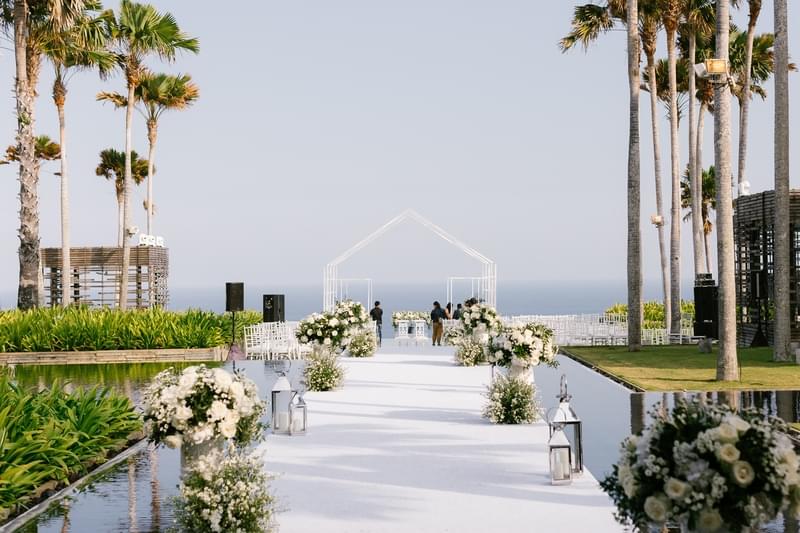  Elegant wedding aisle setup at Alila Villas Uluwatu, featuring white floral arrangements, lanterns, and a minimalist geometric altar overlooking the ocean under clear skies. 
