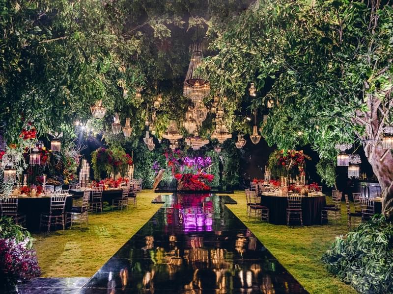  Enchanting outdoor wedding reception at Alila Villas Uluwatu, featuring a reflective black aisle, dramatic crystal chandeliers hanging from lush greenery, vibrant red floral arrangements, and elegantly set tables 