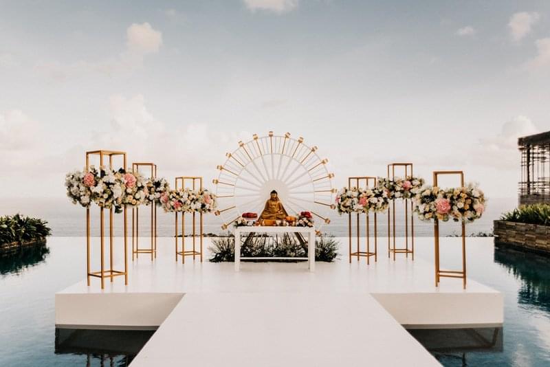 Floating wedding altar setup at Alila Villas Uluwatu, featuring a white stage over the infinity pool, gold frame floral arrangements, and a golden Buddha centerpiece with an ocean backdrop 