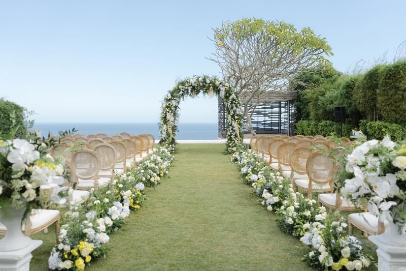  Wedding ceremony setup on the cliff lawn next to the cabana at Alila Villas Uluwatu, featuring an ocean-view backdrop, a floral arch, wooden rattan chairs, and elegant aisle decorations 