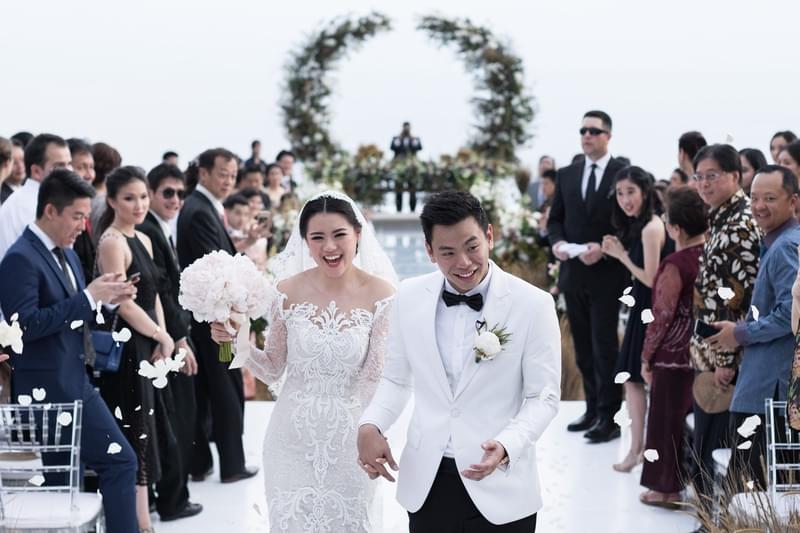  Bride and groom joyfully walking down the aisle after their ceremony at Alila Villas Uluwatu, surrounded by cheering guests and a floral arch in the background 