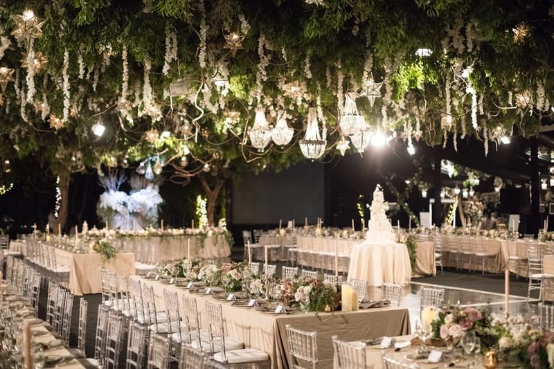  Romantic wedding reception under a marquee at Alila Villas Uluwatu, featuring long banquet tables with elegant floral arrangements, crystal chandeliers, cascading greenery, and a multi-tiered wedding cake centerpiece 
