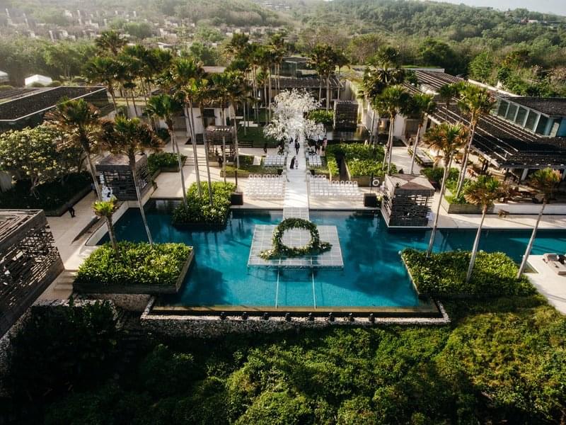  Aerial view of a wedding ceremony on a floating stage over the central infinity pool at Alila Villas Uluwatu, surrounded by lush greenery and perched on a dramatic cliff edge 