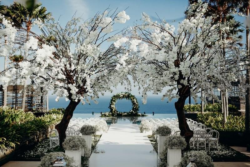  Elegant wedding aisle framed by blooming white floral arches, leading to a floating stage with an ocean-view backdrop at Alila Villas Uluwatu. 