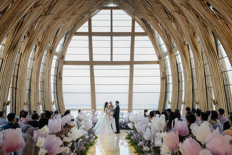 Bridal couple exchanging vows in the Apurva Chapel with large windows and ocean view, surrounded by floral arrangements at The Apurva Kempinski, Bali. 