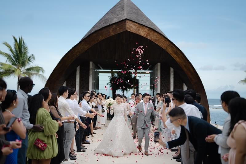  Newlywed couple exiting The Apurva Kempinski’s Majestic Chapel with rose petals being thrown by guests in celebration, with ocean views in the background 
