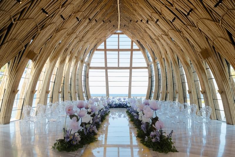  Inside view of The Apurva Kempinski's Majestic Chapel, with a flower-lined aisle, clear chairs, and large windows offering a scenic ocean view 