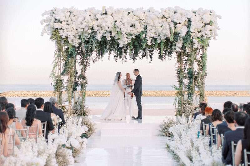  Beachfront Lawn wedding venue at The Apurva Kempinski with bride and groom taking thier marriage vows standing under a floral arch, with the ocean in the background 