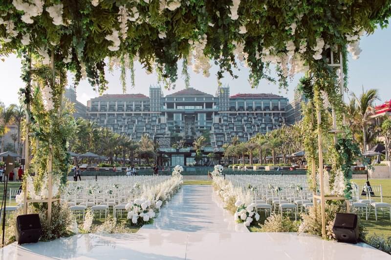  Wedding ceremony setup at The Apurva Kempinski, with a floral arch framing the aisle leading to the resort, white chairs, and a scenic view of the hotel's facade 