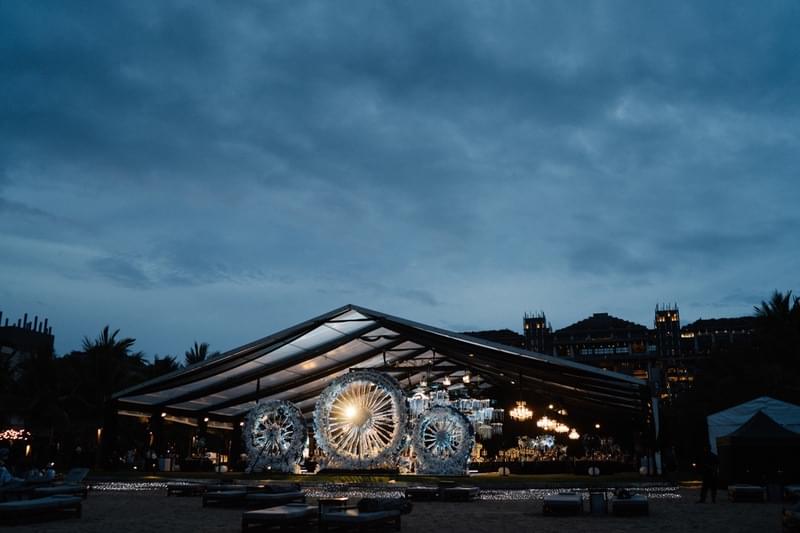  Illuminated wedding reception setup under a clear tent at The Apurva Kempinski Bali's beachfront lawn, featuring intricate lighting and decorative elements against a dusky evening sky 
