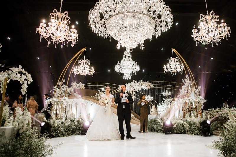  Bride and groom on a glamorous stage under sparkling chandeliers in the Candi Ballroom at The Apurva Kempinski, surrounded by elegant floral arrangements and golden decor 