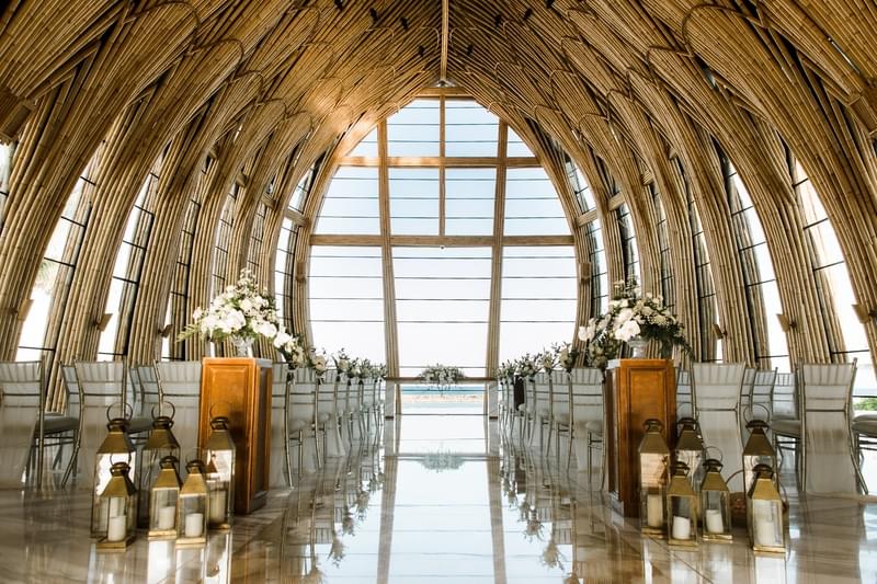  Inside view of The Apurva Kempinski’s Majestic Chapel, featuring bamboo architecture, elegant flower arrangements along the aisle, and large windows with a scenic ocean view 