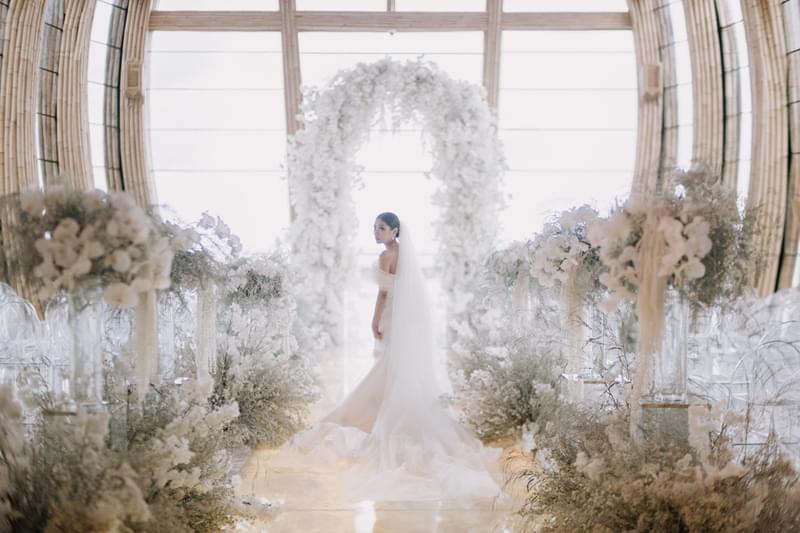  Bride posing at the end of the aisle inside the Majestic Chapel at The Apurva Kempinski, surrounded by white floral arrangements with a beautiful natural light backdrop 
