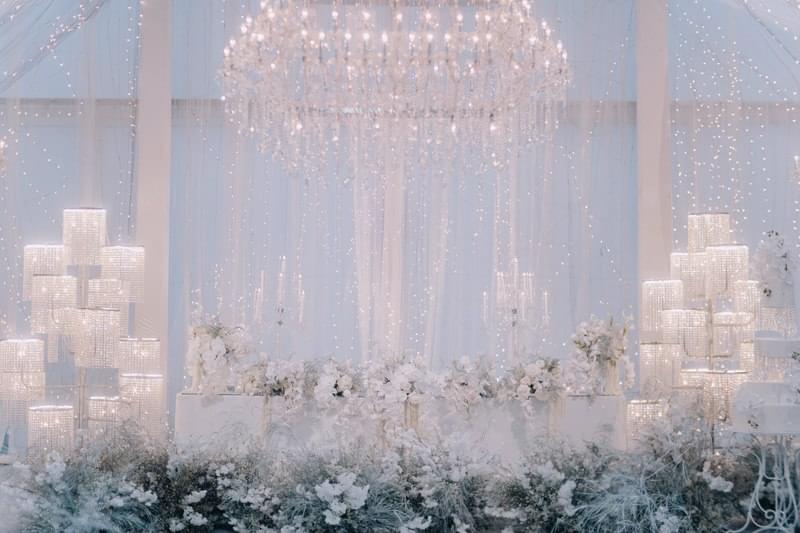  Luxurious bridal table adorned with white floral arrangements, shimmering chandeliers, and cascading lights, creating an ethereal atmosphere in the Candi Ballroom at The Apurva Kempinski 