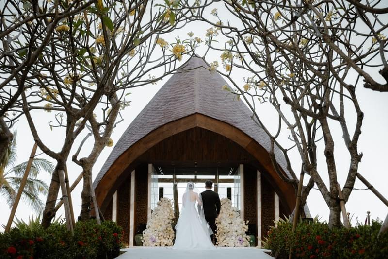  Father walking the bride down the aisle towards the Majestic Chapel at The Apurva Kempinski Nusa Dua, framed by blooming trees and a flower-decorated entrance. 