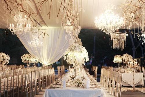  Luxurious wedding reception with crystal chandeliers, white floral centerpieces, and rustic branch decor under a draped canopy.