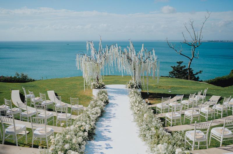  edding ceremony setup at Ayana Sky Amphitheatre with white floral aisle arrangements, delicate hanging blooms on tree structures, and a breathtaking ocean view backdrop. 