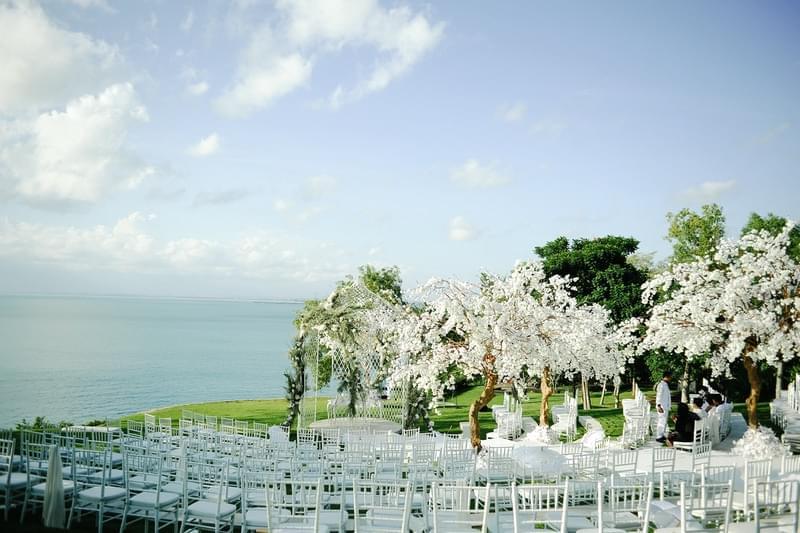  Beautiful outdoor wedding setup at Ayana Sky Amphitheatre with white chairs and blossom-filled trees, overlooking the ocean under a clear sky. 