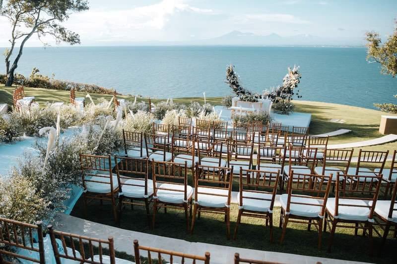  Charming wedding ceremony setup at Ayana Sky Amphitheatre with natural wooden chairs, delicate greenery decor, and a stunning ocean and mountain view backdrop. 
