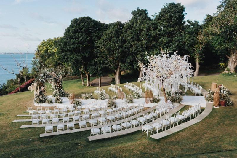  Stunning wedding ceremony layout at Ayana Sky Amphitheatre with curved rows of white chairs, lush floral arrangements, and a backdrop of greenery and ocean views 
