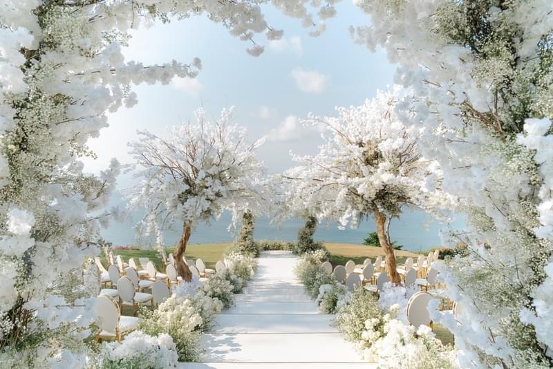  Elegant wedding ceremony setup at Ayana Sky Amphitheatre featuring lush white blossoms on trees lining the aisle, with ocean views in the background. 