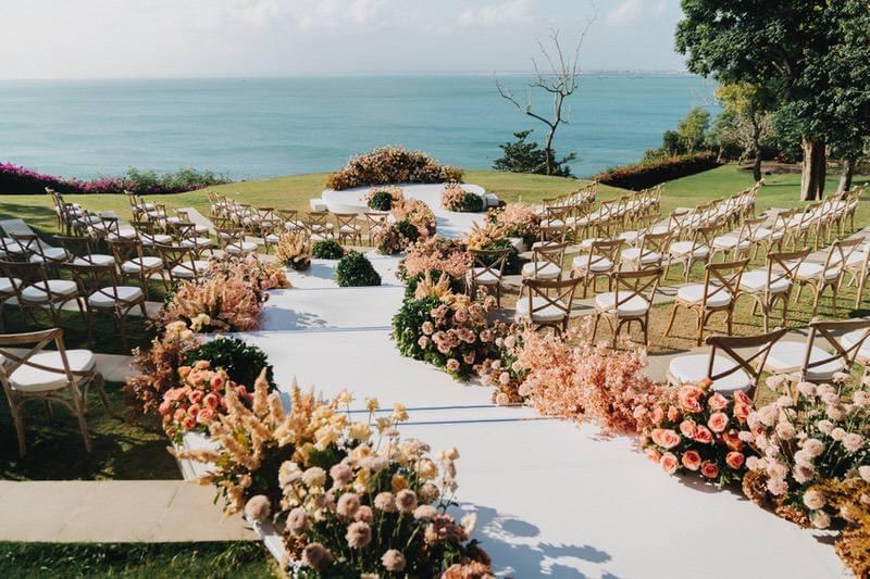  Romantic wedding ceremony setup at Ayana Sky Amphitheatre featuring vibrant floral arrangements in soft pink and peach hues, wooden chairs, and panoramic ocean views. 