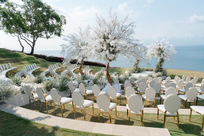  Luxurious wedding ceremony setup at Ayana Sky Amphitheatre with circular seating arrangement, white blossom trees, and a stunning ocean backdrop. 