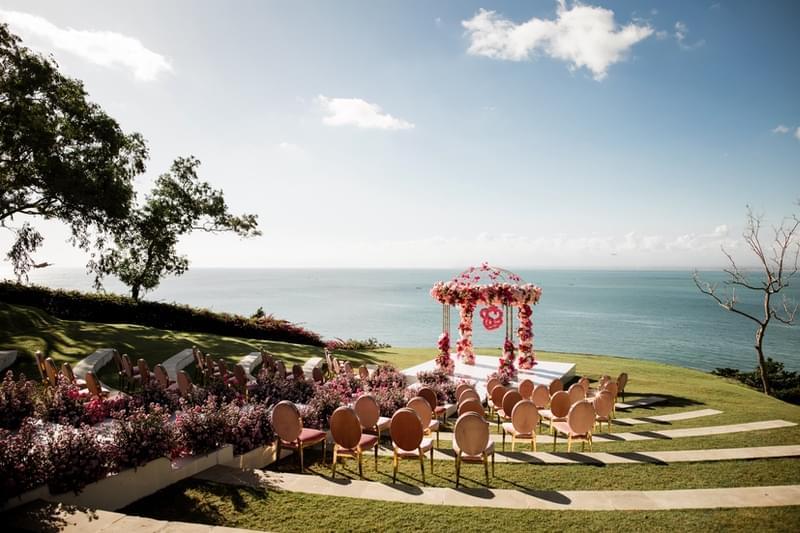  Vibrant wedding setup at Ayana Sky Amphitheatre featuring a pink and red floral mandap with round-back chairs, overlooking the ocean under a clear sky. 
