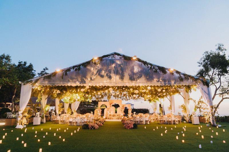  Enchanting evening wedding reception in a draped marquee with hanging floral arrangements, soft candle lighting, and an open lawn setting. 