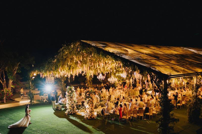  Nighttime wedding reception under a grand marquee with lush greenery, floral arrangements, and crystal chandeliers, as the bride and groom make their entrance on the lawn. 