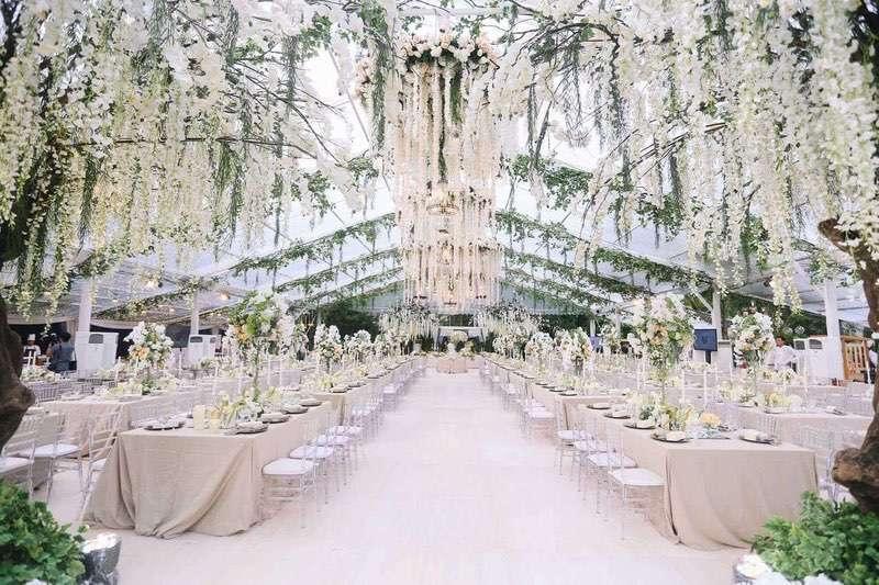  Elegant wedding reception with cascading white floral ceiling installations, long banquet tables, and transparent chairs under a glass marquee. 