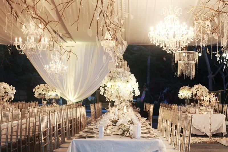  Luxurious wedding reception with crystal chandeliers, white floral centerpieces, and rustic branch decor under a draped canopy.