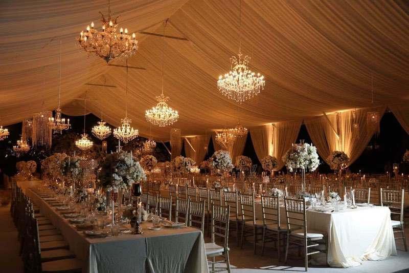  Elegant wedding reception under a draped marquee with crystal chandeliers, gold chairs, and lavish floral centerpieces, creating a warm and luxurious atmosphere. 