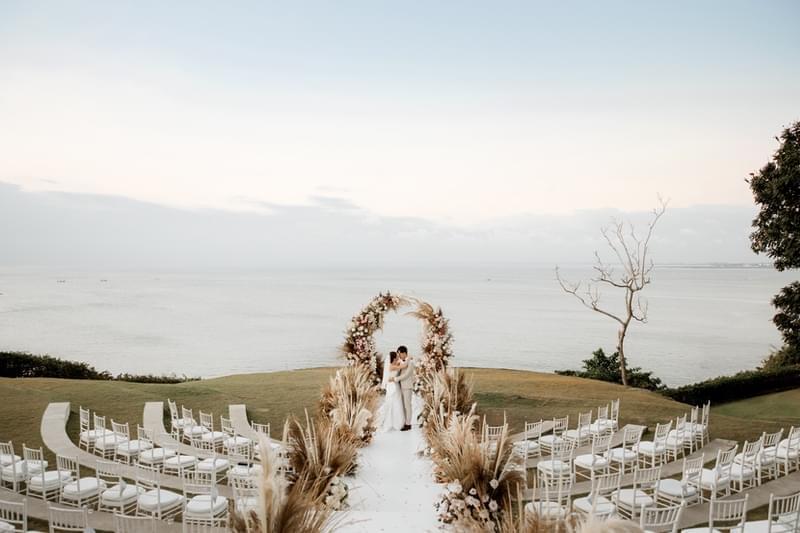  Romantic wedding ceremony at Ayana Sky Amphitheatre with a couple embracing under a floral arch, surrounded by white chairs and pampas grass decor, overlooking the ocean at sunset 