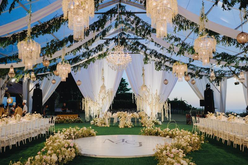  Luxurious wedding reception under a clear tent with hanging chandeliers, greenery, floral decor, and a monogrammed dance floor, creating an elegant outdoor ambiance. 