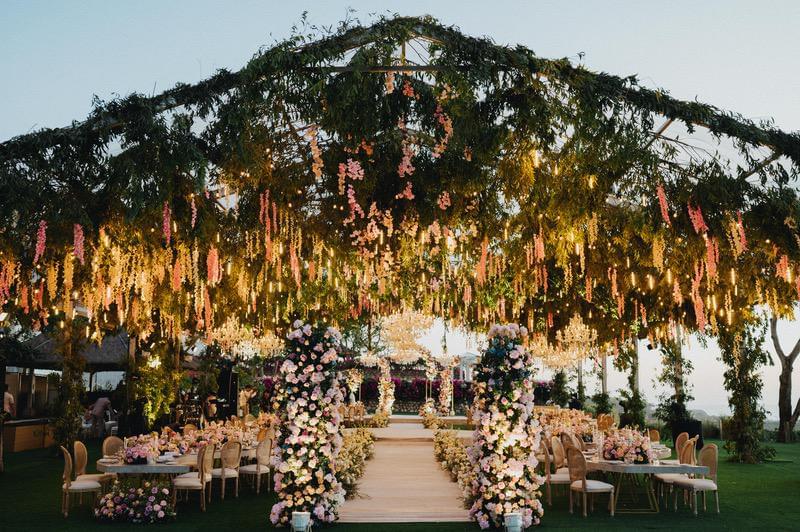  Romantic wedding reception on Ayana’s Middle Lawn with a lush floral canopy of greenery and pink flowers, flower-lined aisle, and elegantly decorated tables under warm ambient lighting. 
