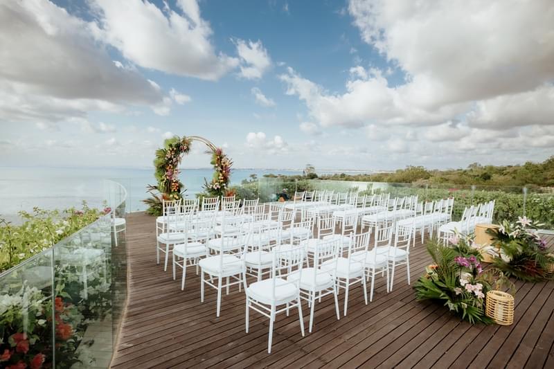 Empty ceremony setup with white chairs and a floral arch on a wooden deck overlooking lush greenery and the ocean at Ayana Villa in Bali.