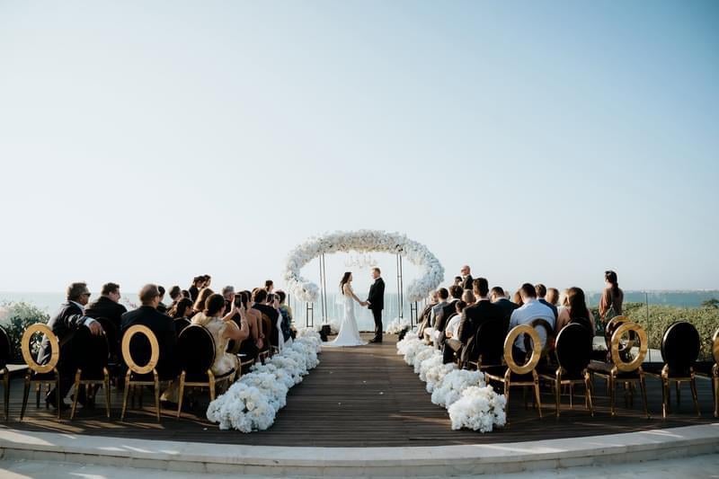 Elegant wedding ceremony at Ayana Villa's clifftop platform, featuring a white floral arch, gold-accented chairs, and panoramic ocean views