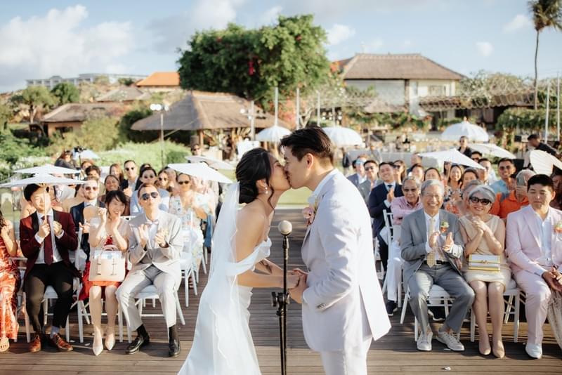 Bride and groom sharing their first kiss as newlyweds during an outdoor wedding ceremony at Ayana Villa, surrounded by joyful guests