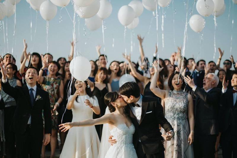 Bride and groom sharing a romantic kiss surrounded by joyful guests releasing white balloons during a celebratory outdoor wedding moment at Ayana Villa