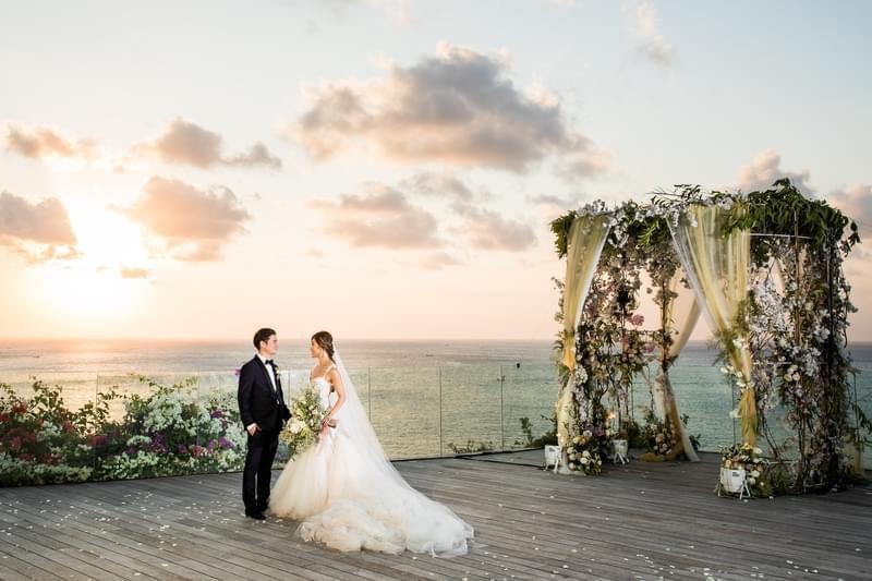 Bride and groom standing on a clifftop deck at Ayana Villa, with a floral wedding arch and a breathtaking ocean sunset as their romantic backdrop