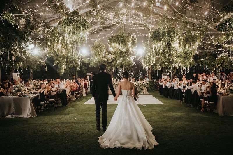 Bride and groom holding hands as they walk onto a beautifully lit dance floor under twinkling fairy lights and lush greenery, surrounded by wedding guests in the Ayana Villa garden