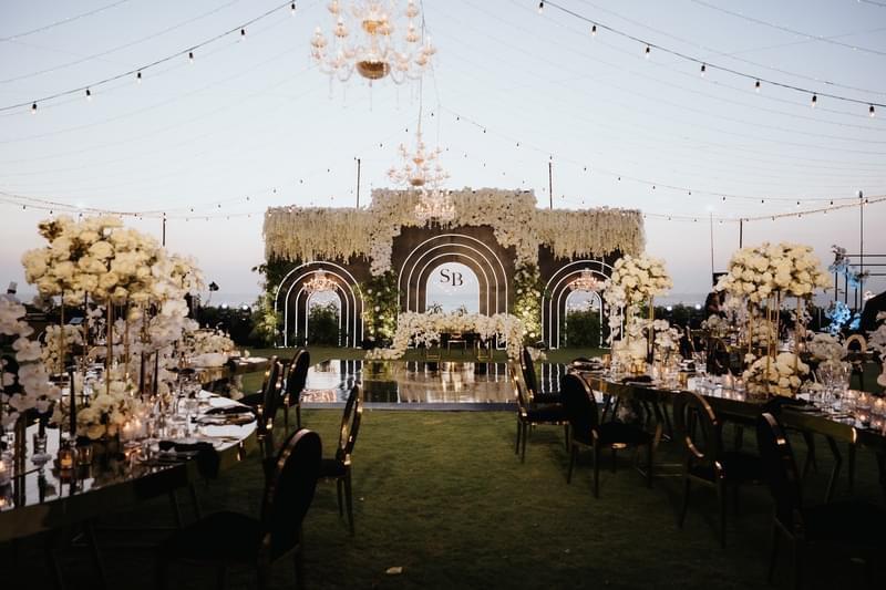 Luxurious garden wedding setup at Ayana Villa featuring a grand floral backdrop, elegant black and gold dining decor, chandeliers, and string lights under the evening sky