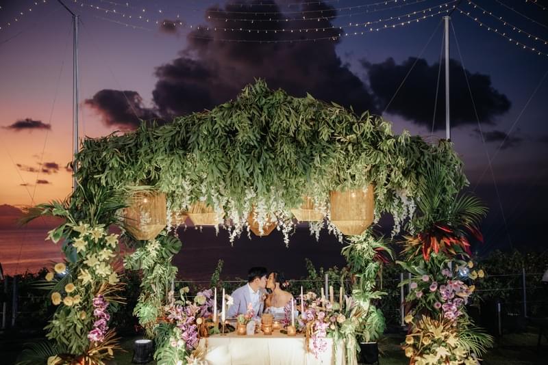Enchanting bridal table adorned with lush greenery and floral accents under a canopy of foliage, set against a romantic sunset backdrop at Ayana Villa